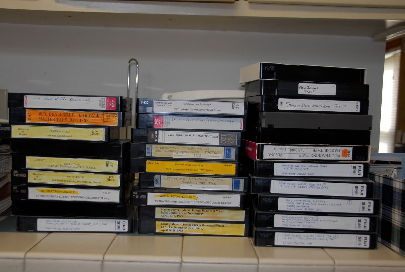 Twenty-nine VHS tapes in three stacks on a shelf. Left: the dealership lab talk master, the Rick Schneider "Document of Record" tapes, Green Team, a Colorado Springs news report and the fifth Ukiah tape. Middle: The Birth of New Technology, the dealership masters, the International Symposium on New Energy workshop and main talk, and two copies of the IANS talk of April 1993. Right: the New Zealand tapes, the WFC teaching tape master of April 1988, and the Ukiah SEER '94 master copies.